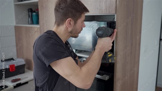 Man in workwear installing built in microwave above oven using cordless screwdriver in wooden kitchen cabinet