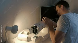 Young repairman master is repairing sewing machine sitting at table, side view. Unscrew body of sewing machine using screwdriver. Man is testing and disassembles the sewing machine.