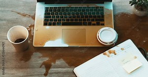 Spilling coffee on a laptop keyboard at work in a modern office from above. Clumsy woman hands at work making a mess at her office desk with cup of liquid falling over equipment at the workplace