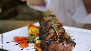 chef in background sprinkles seasoning slow motion. Chef adds a pinch of seasoning to the cooked and plated steak. The camera circles in slow motion around the steak and steamed vegetables on a white