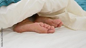 Close-up image of adult bare feet under white bed sheets, suggesting relaxation or sleep in a bedroom.