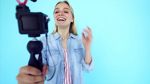 Vlogger uses camera and selfie stick to record video blog in front of a blue background