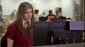 A nurse works on a computer in a busy hospital nurses station with a focused and neutral expression.