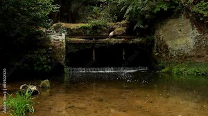 Small weir on the river flows out from cave. Cold water of small river flow over small stony weir. Stony and rusty construction of weir.