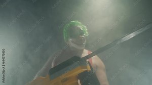 Scary man in the horror clown mask with green hair, standing in suspenders in the dark studio with flashing lights and threatening with the chainsaw close up