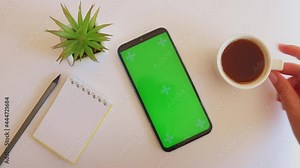 Work desk with notepad, pencil, plant and green screen smartphone. Woman's hand puts coffee on the work table
