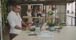 Senior male manager working alone at his desk while his colleagues are working together in the background in the office during summer time