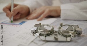Optometry glasses on table and doctor ophthalmic hands filling medical card with pen, cropped view. Ophthalmologist visit concept, eyesight check up