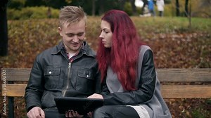 Young woman with red hair and attractive man in a leather jacket sitting on a bench in park and using a digital talbe while discussing someting. They are choosing a trip and shopping online