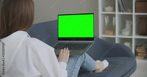 Woman at Home Sitting on a Couch Works on a Laptop Computer with Green Mock-up Screen. Coronavirus Covid-19 quarantine remote education or working concept. Girl Using Computer, Browsing through