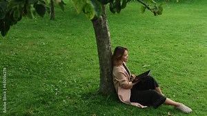 Young Asian woman sitting under tree in park and working using laptop computer. Female millennial relaxing outdoors, browsing Internet, checking news feed or drawing with a tablet