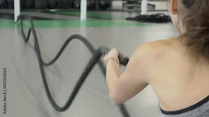 Young woman doing exercise with rope in sports club. Female stands, holds strong cord in hands and quickly moves it across floor, making double waves