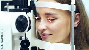 Face of a young female at an ophthalmologist office. Smiling woman holds eyes open during an optical exam. Eye check up by modern microscope. Medical device laser shine inside the eye.
