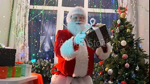 Active Santa Claus dances with colorful gift boxes that fly into his hands and putting them on table in a large mountain of presents, having fun near Christmas tree and colorful garlands on Xmas party
