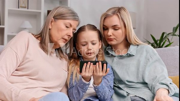 Grayhaired woman grandmother, little granddaughter and blonde mother sit in the living room watching a video on mother's day. A cute girl explains to her family how to use programs on the phone.