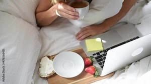 Woman take cup and drink of coffee. Morning breakfast in bed. Delicious, aromatic coffee, sandwich and two ripe strawberries on wooden board. Work on laptop. home-office in time quarantine
