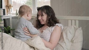 One Caucasian brunette mother in nightwear waking up with her tiny newborn, playing on bed. Young woman with baby relaxing at home. Bed-sharing concept. Infant child with his mom in bedroom.
