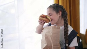 schoolgirl child eating hamburger breakfast at recess. training nutrition for schoolchildren fast food concept. schoolgirl child eating breakfast sandwich from a paper bag with a backpack lifestyle