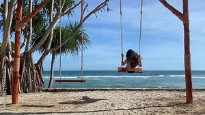Young woman sitting on swing on the beach on blue sea and green palms background and having fun.