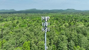 Cell tower with 4G/5G antennas high above the tree tops in a forested area of northeast United States on a summer day. Aerial drone shot orbiting the wireless telecommunications tower.