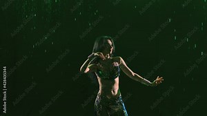 Charming oriental dancer passionately dances an exotic dance in revealing shiny costume against background of falling drops of rain, smoke and green light. Close up. Silhouette. Slow motion.