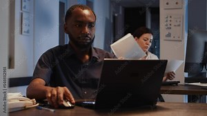 African american man working on laptop in startup office, analyzing data on internet and documents late at night. Using research to create report and presentation with app. Handheld shot.