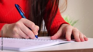 A girl at the table writes with a pen in a notebook. Student studying, taking notes. School and education. Closeup video motion footage
