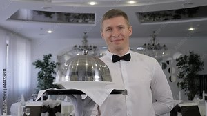 A male waiter holds a tray with a covered dish in a restaurant.