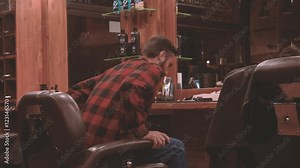 Lumberjack sits down on barber chair in barbershop HD video. Bearded man in vintage authentic hairdressing salon