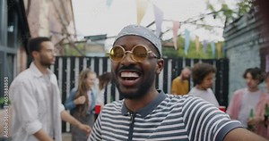 Overjoyed African American man making online video call at open air party speaking showing dancing friends toasting. Communication and modern lifestyle concept.