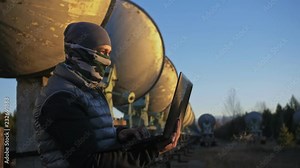 Hacker hacks the radio telescope using notebook. Man to program hacking on laptop in nature. Terrorist is hiding in camouflage balaclava. Hacker has criminal cases on the Internet. Programmer uses a