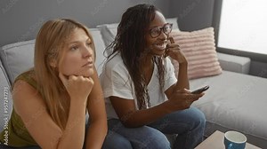 Young women friends watching tv together at home in the living room during the day, one woman happy and the other woman thoughtful