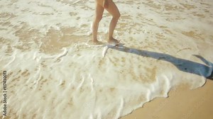 Beautiful bare feet on the beach. Sea foam washes female legs. Image of feet in the sand on the shore in the wave. Sunshine on the ocean in the wave. Summertime holidays Sea waves and sand.