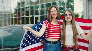 Two beautiful girls stands with an American flag against the background of a classic American car. American patriot women. A girls celebrates Independence Day of the United States of America