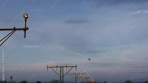 Small private jet flying low over runway approach lights at dawn, with airport control tower silhouetted against the sky