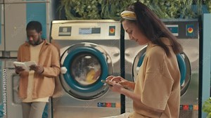 Side tilt footage of young cheerful Caucasian girl sitting on table and chatting on smartphone while waiting for her clothes to finish washing in self-service laundry