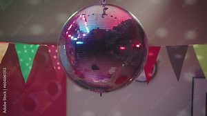A silvery mirrored disco ball rotates and throws multicolored highlights on the walls close up. Party with disco ball and paper flags. Party, birthday, celebration, disco.