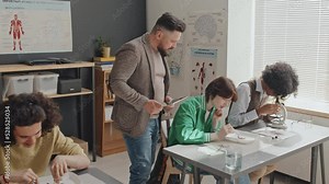 Medium long of bearded male teacher walking in classroom, watching diverse high school students sitting at desks, using microscopes and dissecting frogs Stock Video