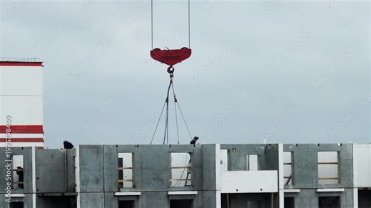 Construction site. Red crane hook over concrete panels of new building. Workers on panels perform installation. Overcast sky. Industry, labor, development.