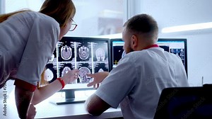 In Control Room Doctor and Radiologist Discuss Diagnosis while Watching Procedure and Monitors Showing Brain Scans Results, In the Background Patient Undergoes MRI or CT Scan Procedure.