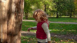 Cute little girl walks in the autumn forest with a tree. Child with curly blond hair in a pink vest on background of the park on a beautiful sunny and warm autumn day. Slow motion. Close up.