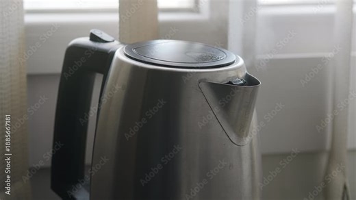 Boiling stainless steel electric kettle placed on a kitchen countertop near a window making steam jets