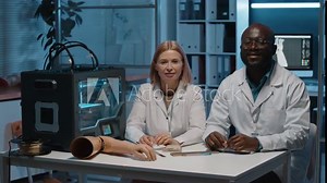 Portrait of two laboratory workers sitting in laboratory next to 3D printing machine and arm prosthetics