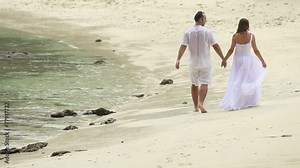 blonde bride and handsome groom walk along sandy beach under rai