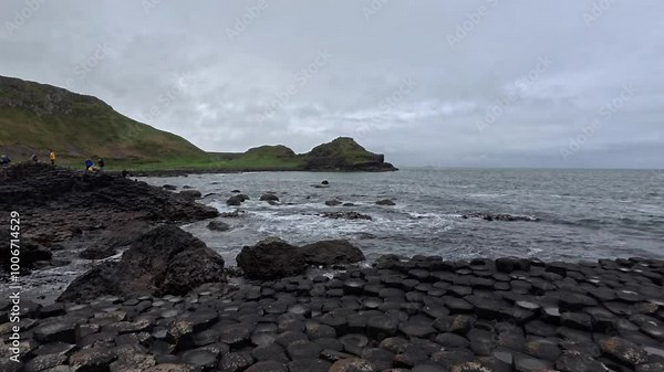 Tourists explore the hexagonal basalt columns at Giant's Causeway, Northern Ireland, with the rugged coastline and green hills in the background. The Atlantic Ocean waves gently roll in