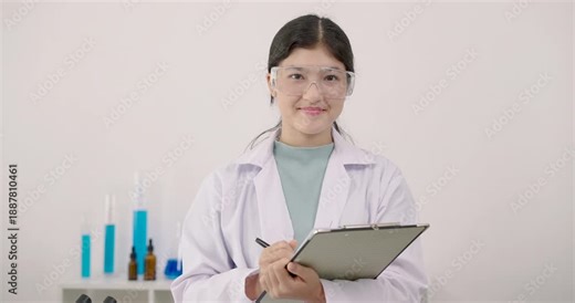 Asian female student writing experiment notes on clipboard with pen during science class in classroom lab wearing labcoat and goggles surrounded by testtubes and colorful beakers on desk