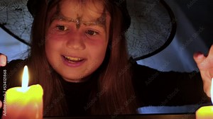 Crazy little witch reading spells at Halloween night by candlelight. Child dressed in costume and hat plays a witch in school play