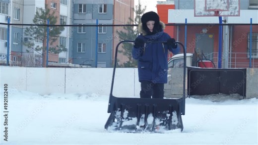 Teenager clearing snow on a sports ground using a large snow pusher. Winter cleaning of the backyard area near residential buildings