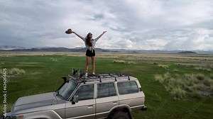 Travelling by car. Young woman shape rising hands standing on the car roof on the sunrise. Enjoying beautiful cloudscape in the morning