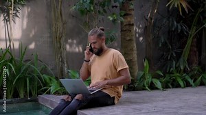 Caucasian male in casual wear using laptop computer for remote job at pool terrace talking on smartphone, skilled man making cellular call using roaming connection during distance job Stock Video